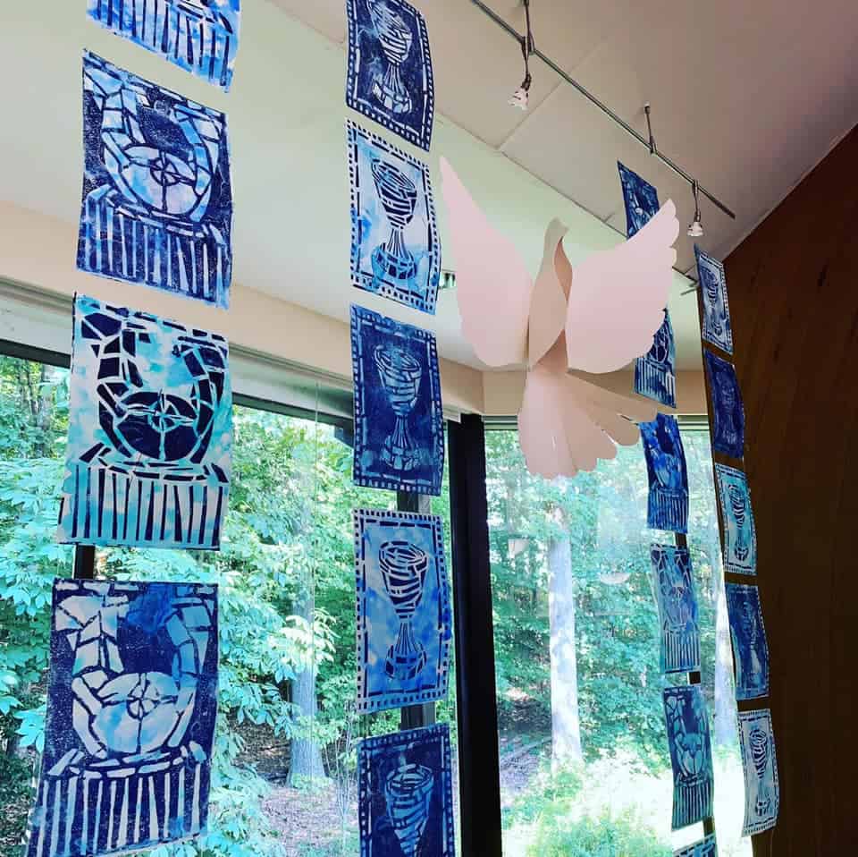 Blue patterned artwork hangs from ceiling by a window, alongside a white paper dove, overlooking a lush green forest.