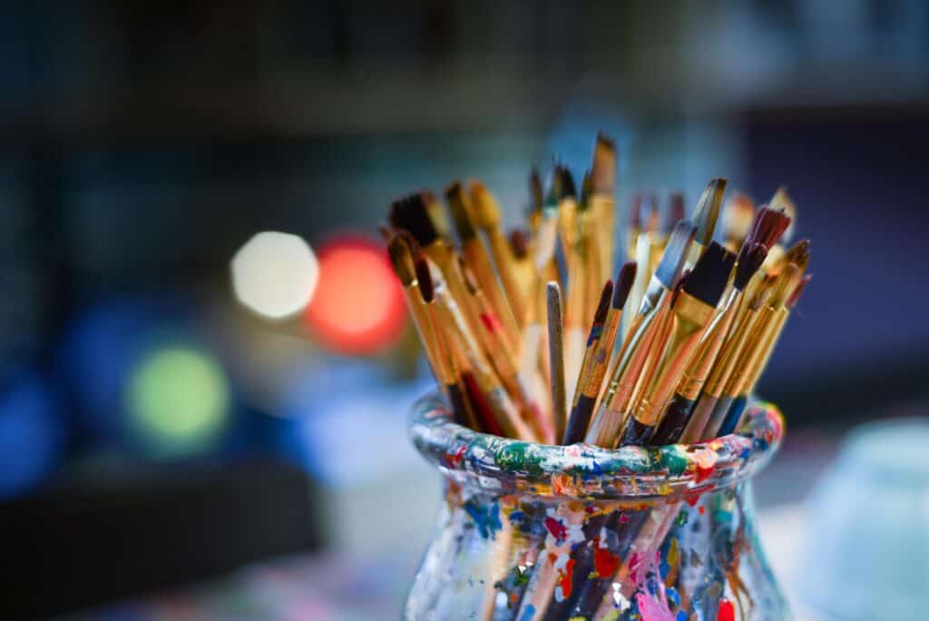 Paintbrushes in a jar with colorful paint splatters for art projects.