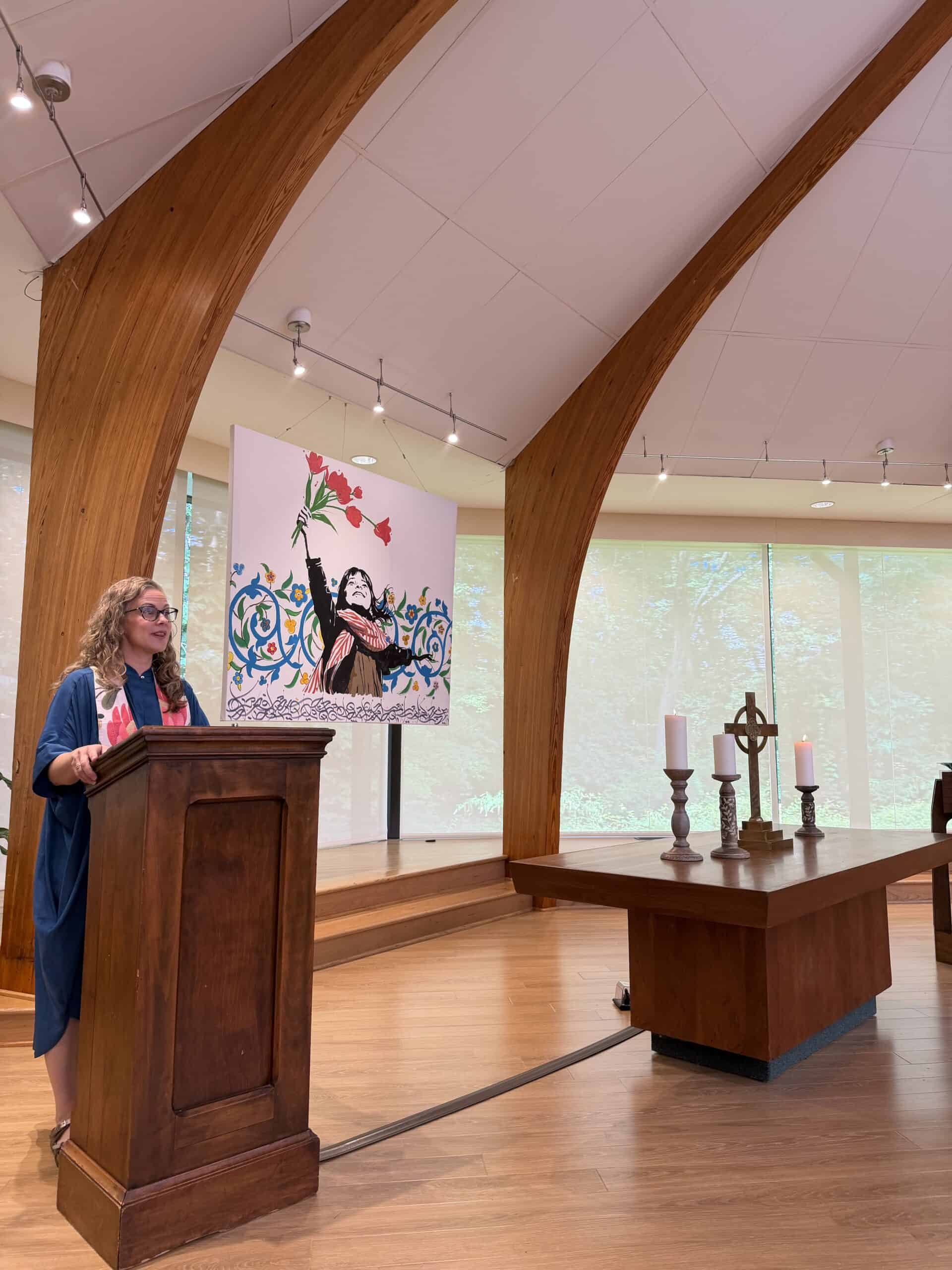 A person speaks at a wooden podium inside an elegantly designed room with large windows, surrounded by colorful flowers and wooden arches.